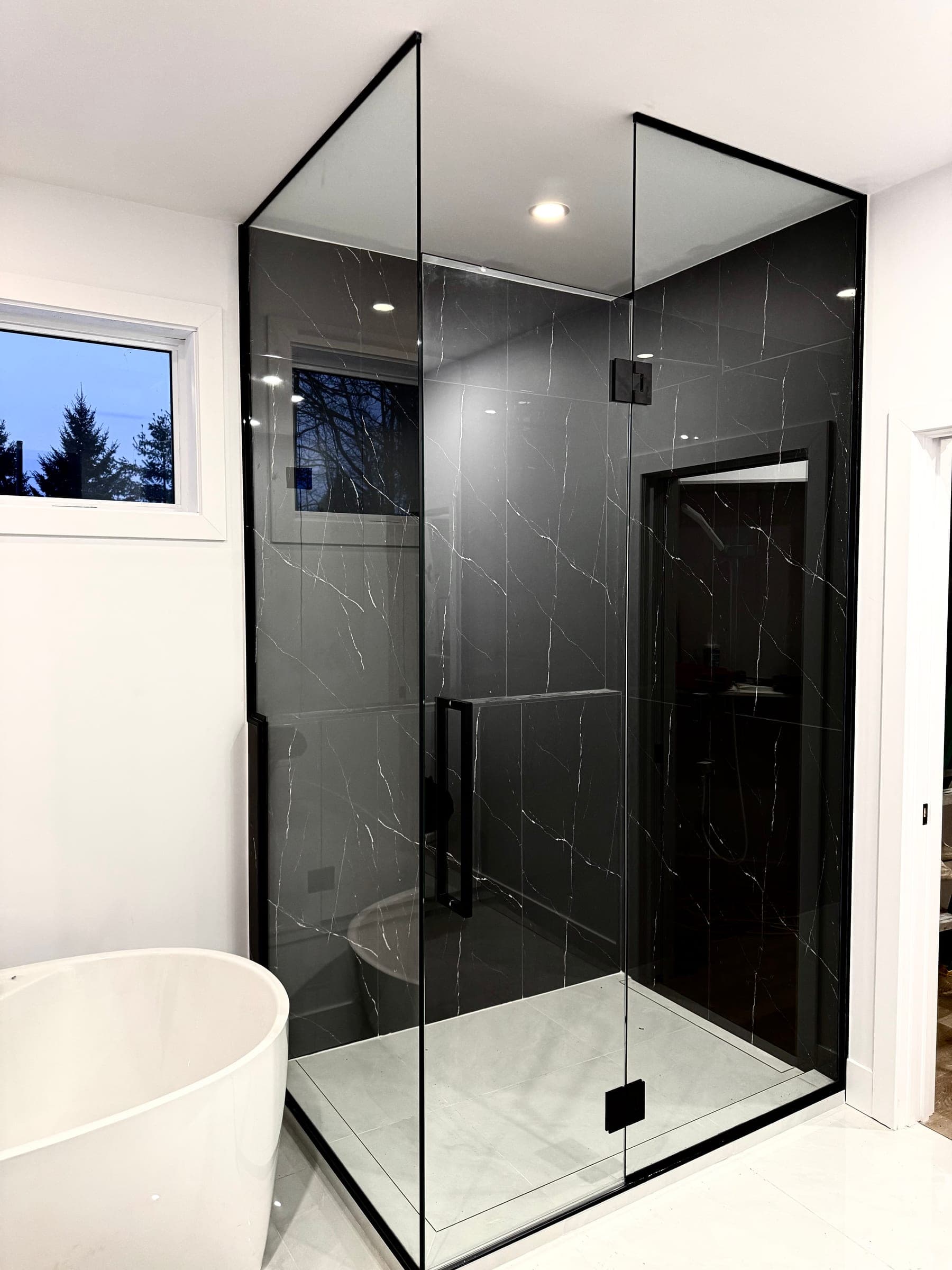 A corner shower with a slim black frame on veined black marble, beside a white freestanding bathtub